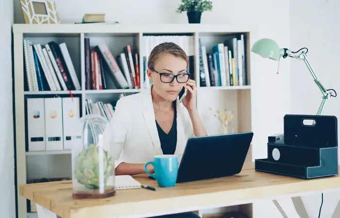 Focused businesswoman in white blazer working on laptop and talking on phone at modern home office desk with bookshelves